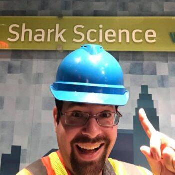 Dr. David Shiffman wears a blue hard hat and points to a sign that says Shark Science