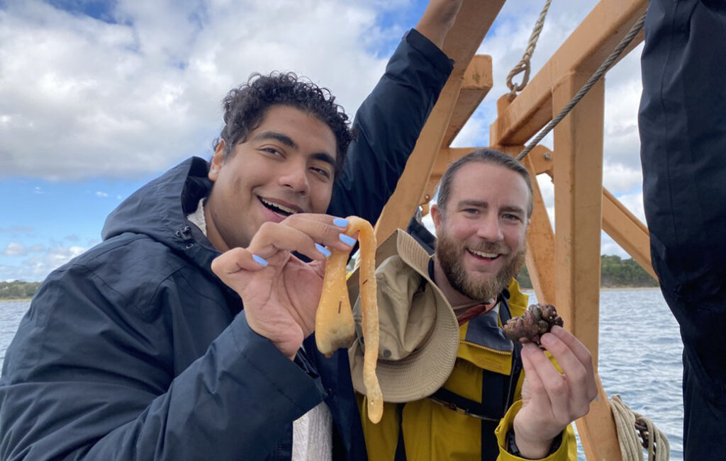 Anil Oza and Bryce Hoye smile and hold up specimens while aboard a boat. 